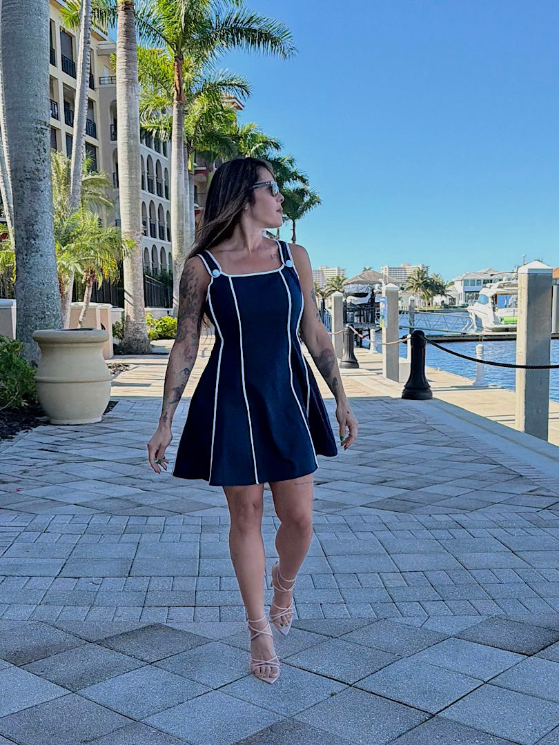 Woman in a navy dress walking along a waterfront promenade with palm trees and buildings in the background.