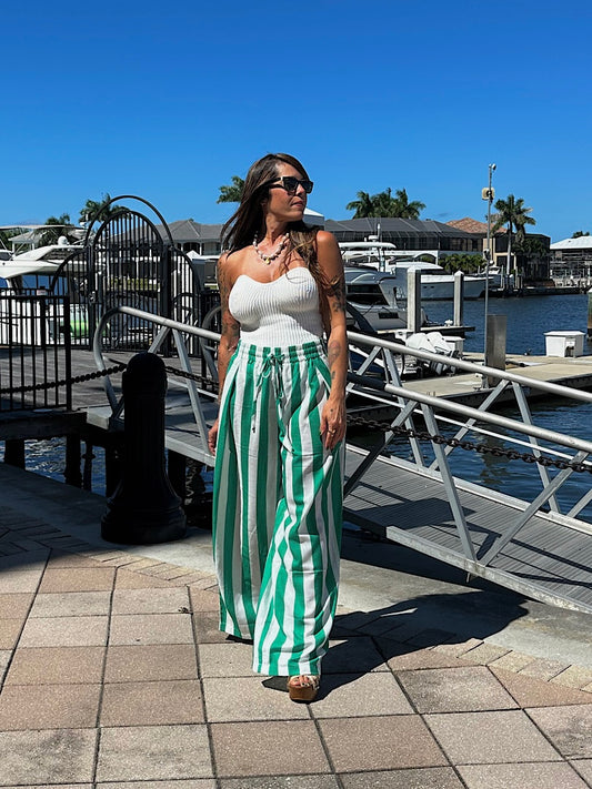 Woman in a white top and green striped pants standing on a dock with boats and palm trees in the background.