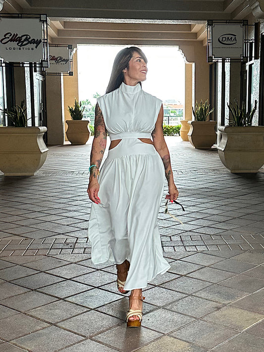 Woman in a white dress walking in an outdoor setting with potted plants and a building in the background.