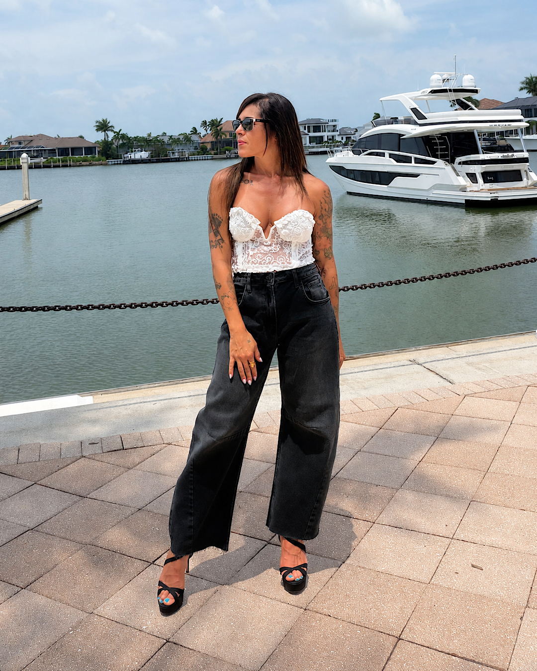 A woman stands on a paved waterfront walkway wearing a white lace bustier-style top, black wide-leg jeans, and black sandals, with water, boats, and a marina in the background.