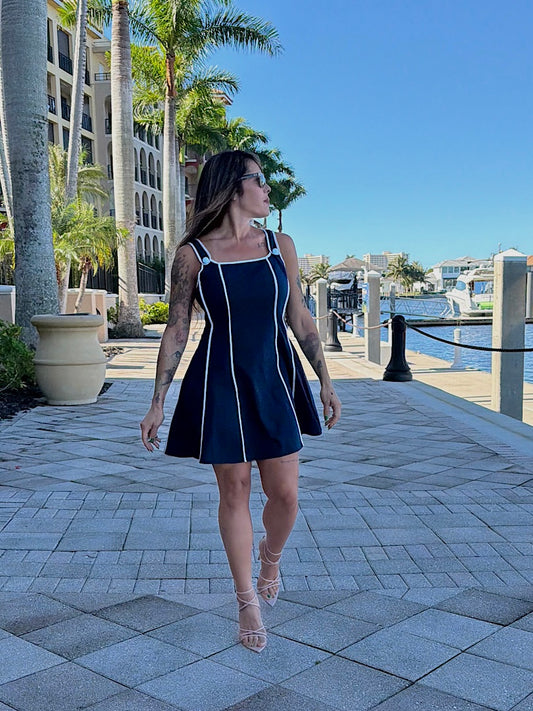 Woman in a navy dress walking along a waterfront promenade with palm trees and buildings in the background.