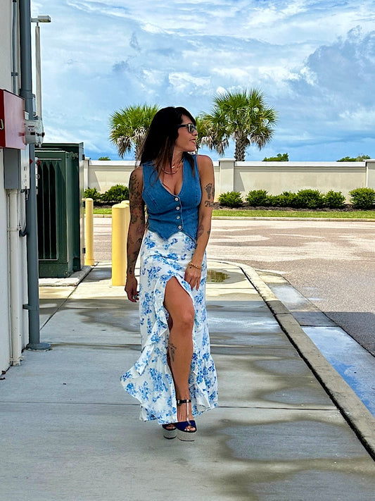 Woman in a blue top and floral skirt standing on a sidewalk with palm trees in the background.