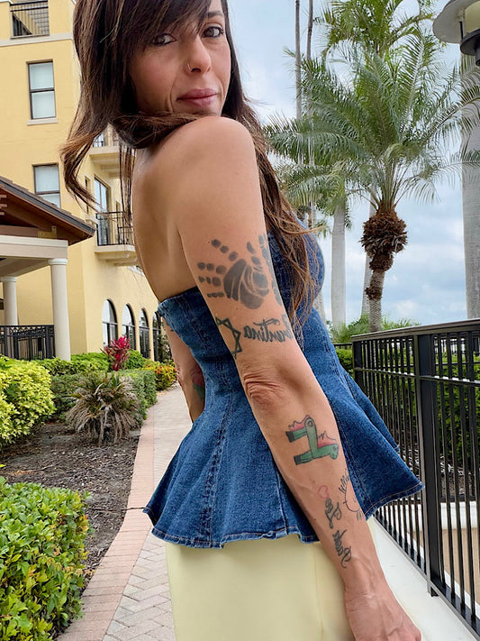 Woman with tattoos on her arms standing outdoors with palm trees and a building in the background