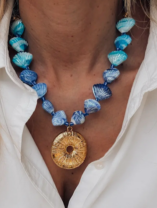 Necklace with blue and white shell-like beads and a gold pendant worn by a person in a white shirt.