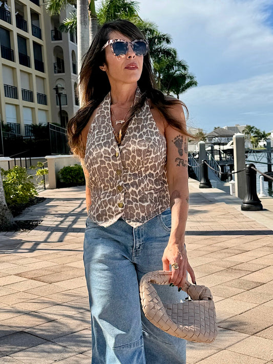 Woman wearing a leopard print top and blue jeans, standing on a sidewalk with buildings and palm trees in the background.