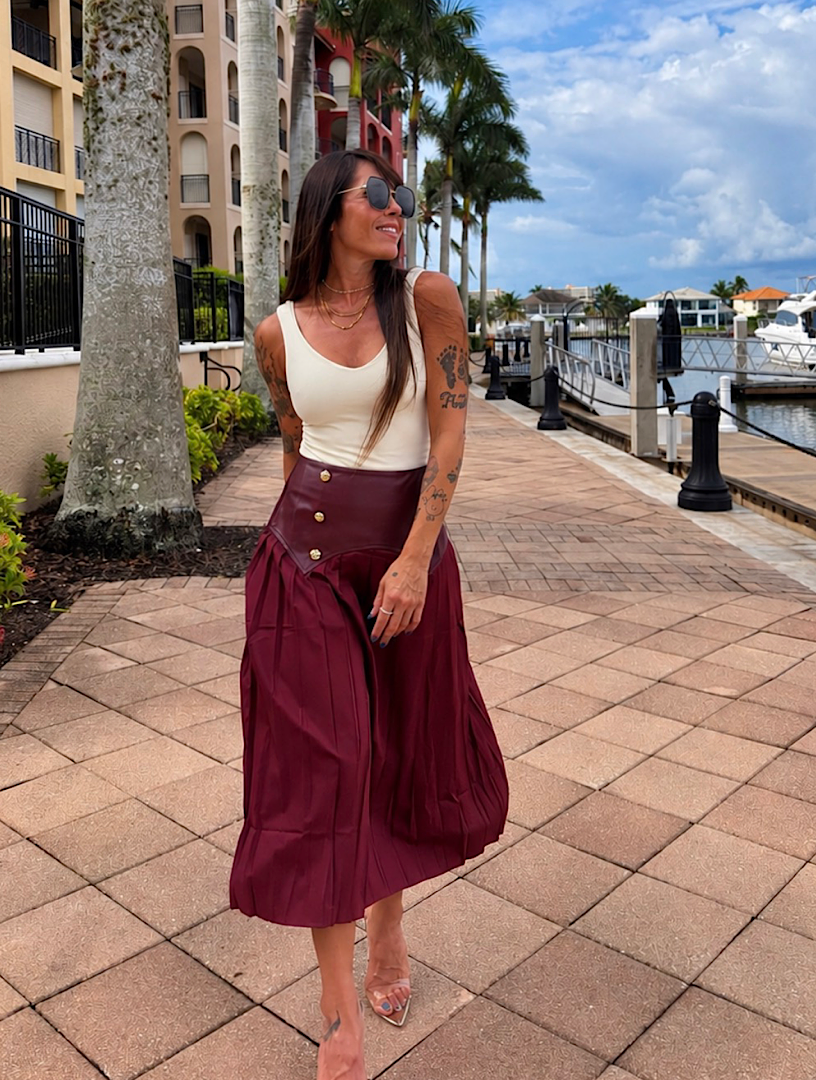A woman stands on a waterfront walkway wearing a sleeveless cream top and a high-waisted, pleated burgundy midi skirt with gold-tone buttons. Palm trees, buildings, and a marina are visible in the background.