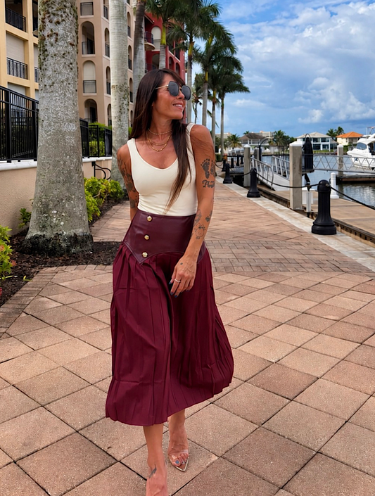 A woman stands on a waterfront walkway wearing a sleeveless cream top and a high-waisted, pleated burgundy midi skirt with gold-tone buttons. Palm trees, buildings, and a marina are visible in the background.