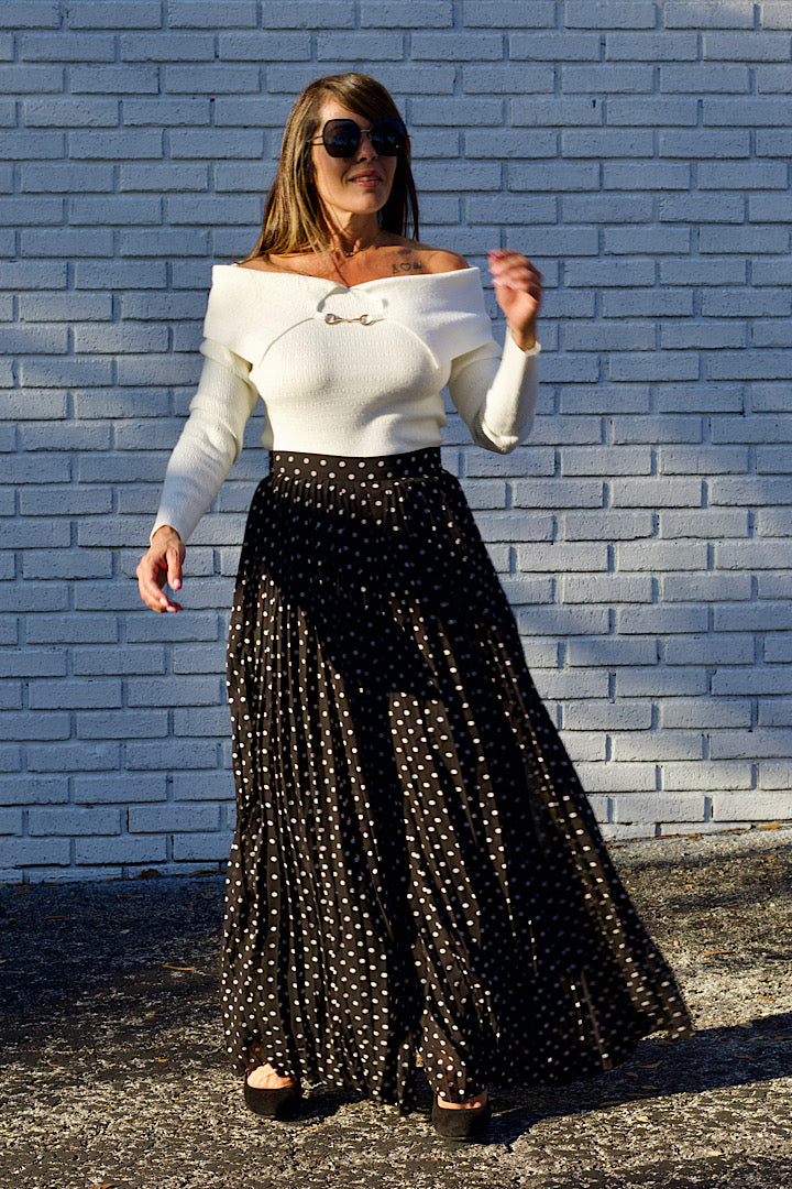 Woman wearing a white off-shoulder top and black polka dot skirt against a light blue brick wall.