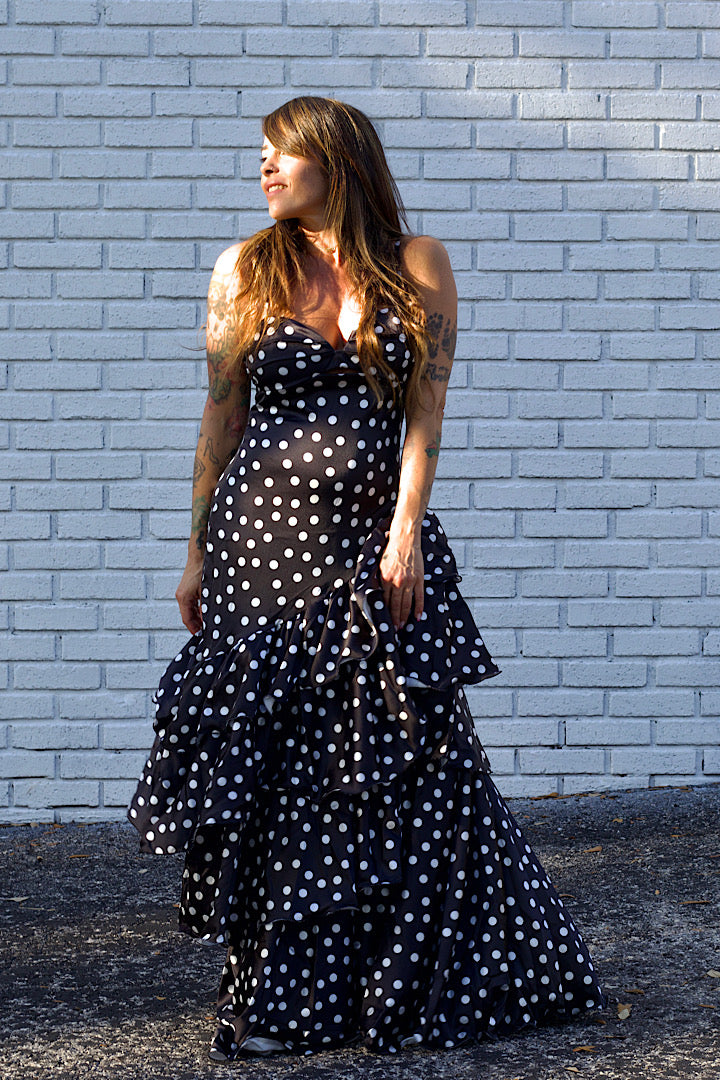 Woman wearing a black polka dot dress against a light gray brick wall.