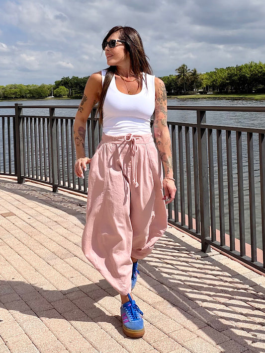 Woman in white tank top and pink pants standing on a wooden boardwalk by a waterfront.