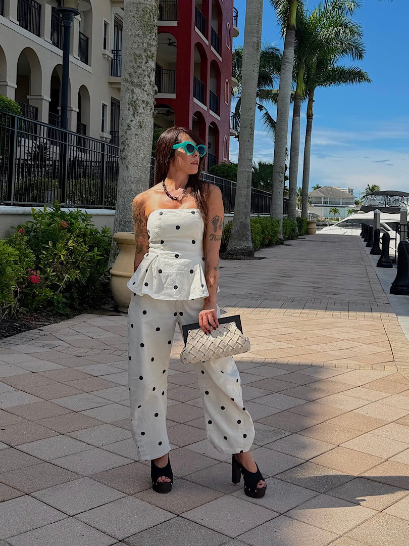 A woman stands on a sunny outdoor walkway wearing a white strapless peplum top and matching wide-leg pants with black polka dots, holding a quilted handbag. Palm trees, buildings, and a blue sky are visible in the background.