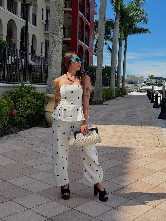 A woman stands on a sunny outdoor walkway wearing a white strapless peplum top and matching wide-leg pants with black polka dots, holding a quilted handbag. Palm trees, buildings, and a blue sky are visible in the background.