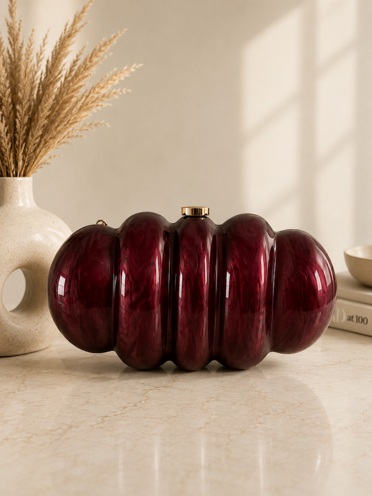A glossy, ribbed burgundy red clutch bag with a small gold clasp, styled on a light marble surface next to a ceramic vase with dried grasses.