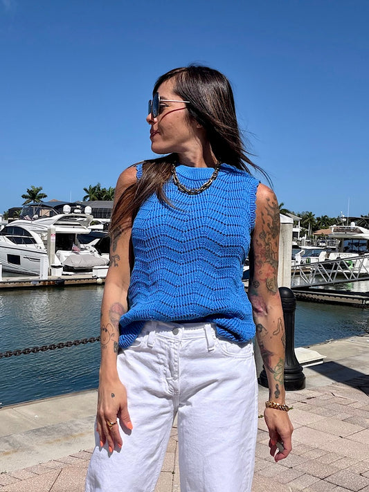 Woman wearing a blue sleeveless top and white pants standing by a marina.