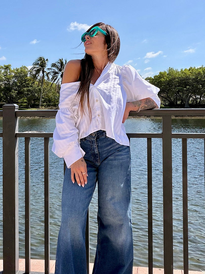 Woman in a white blouse and blue jeans standing by a lake with palm trees in the background
