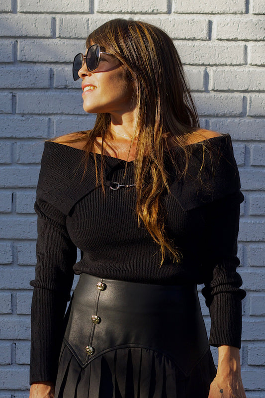 Woman wearing a black off-shoulder top and leather skirt against a gray brick wall.