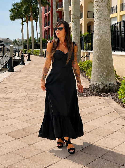 A woman wearing a sleeveless black maxi dress and black strappy heels walks along a paved outdoor walkway lined with palm trees and resort-style buildings.