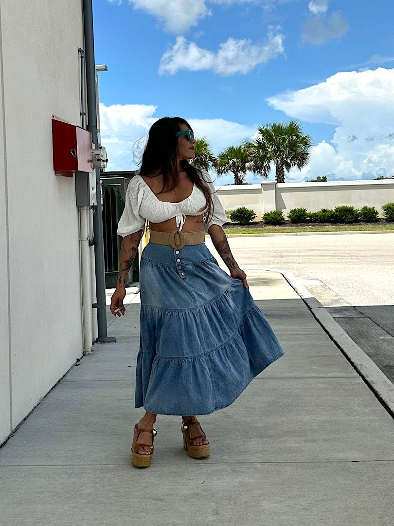 Woman in a white top and blue denim skirt standing on a sidewalk with palm trees in the background.