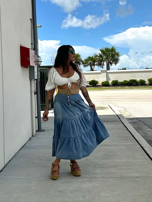 Woman in a white top and blue denim skirt standing on a sidewalk with palm trees in the background.