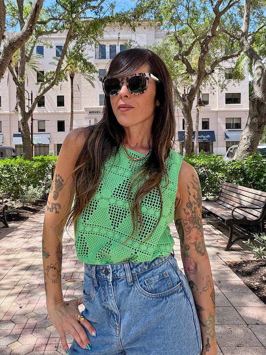 Woman wearing a green top and blue jeans standing outdoors with trees and buildings in the background.