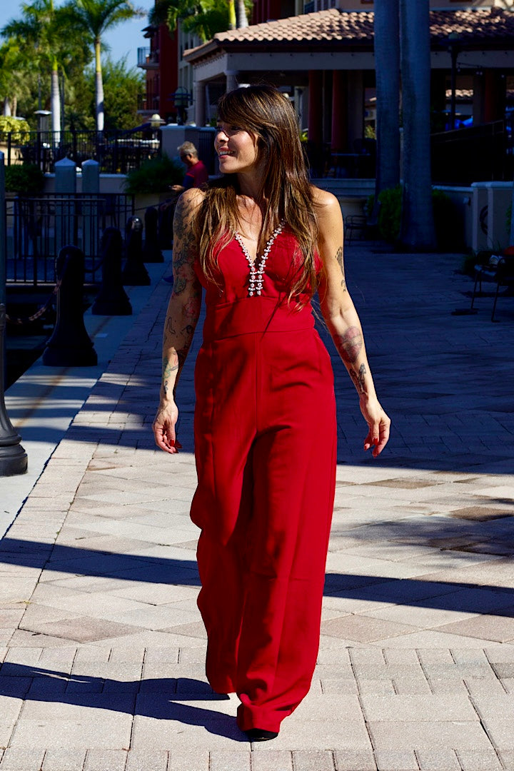 Woman in a red jumpsuit walking on a sidewalk with palm trees and buildings in the background.